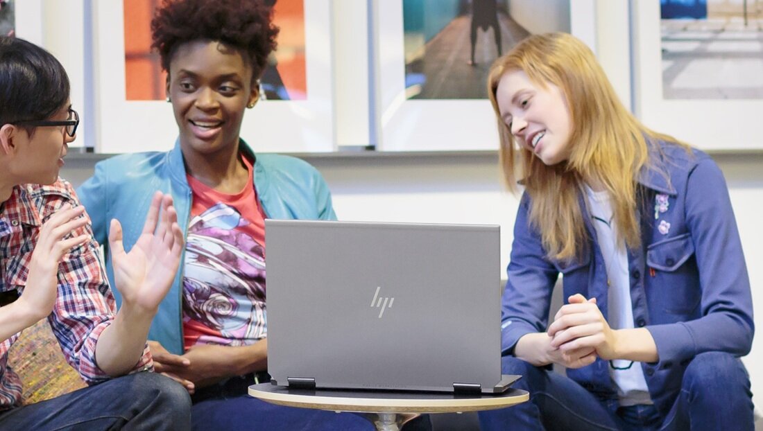 Laptop HP EliteBook 665 G11 na drewnianym stole w kawiarni podczas wideokonferencji, kobieta w czerwonej bluzce na ekranie, obok kolorowe kubki wielokrotnego użytku i roślina doniczkowa, atmosfera pracy zdalnej.