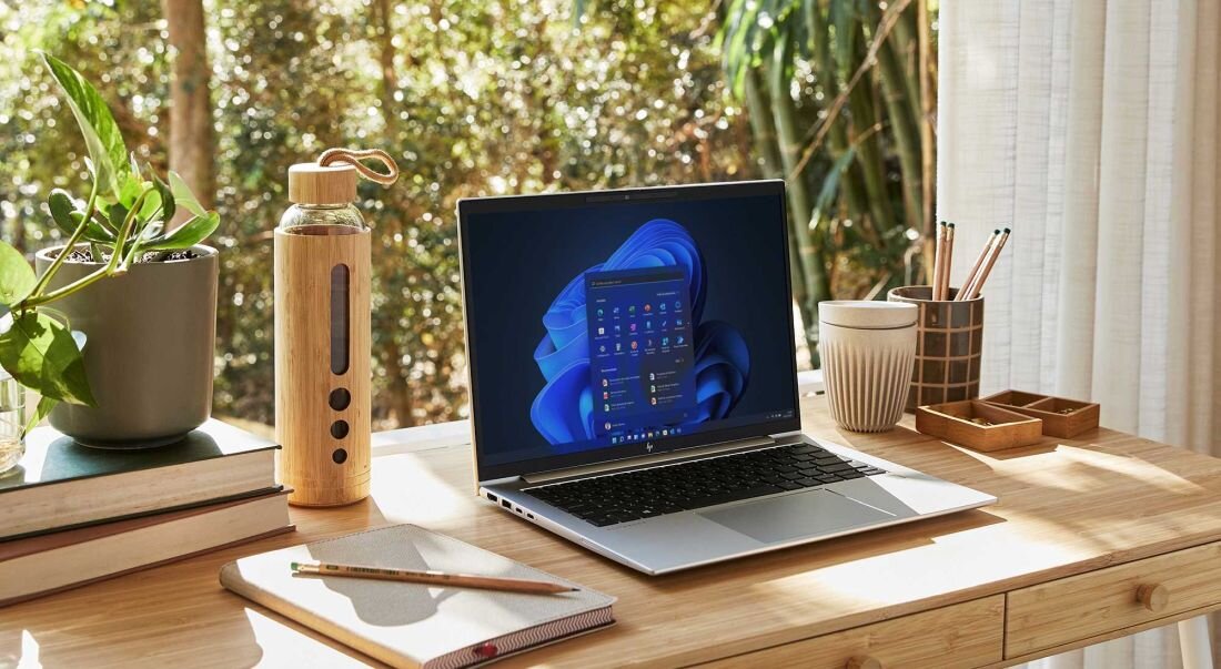 Laptop HP EliteBook 665 G11 na drewnianym biurku z systemem Windows 11, niebieskie tło ekranu z logo Windows, obok bambusowa butelka na wodę, roślina doniczkowa i drewniane akcesoria biurowe, podkreślający ekologiczne podejście i niski TCO. - Certyfikat TCO, Ekologia