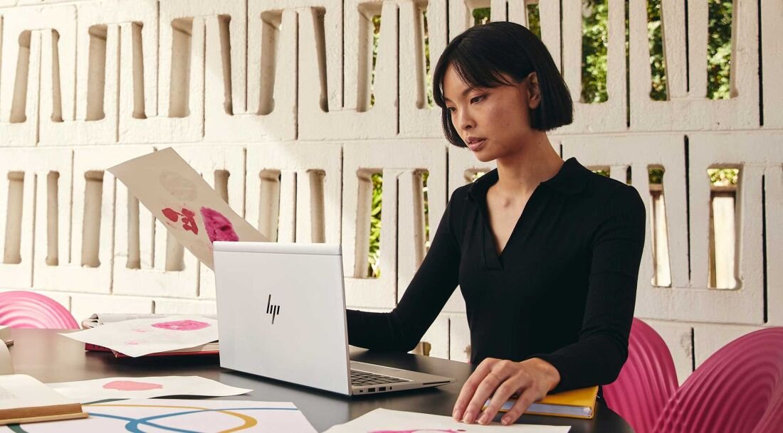 Kobieta w czarnej bluzce pracująca na laptopie HP EliteBook 665 G11 w jasnym wnętrzu, na stole dokumenty z kolorowymi wykresami i diagramami, w tle białe krzesła i wnętrze biurowe. - Bateria 3-komorowa litowo-jonowa o pojemności 56 Wh