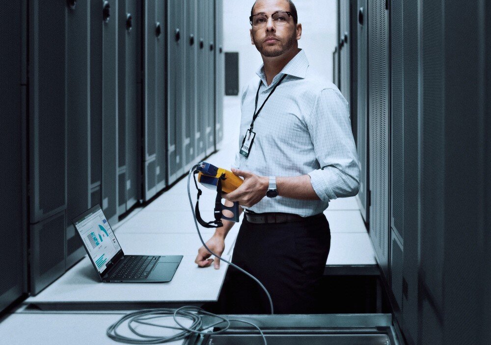 Laptop HP ProBook 440 G11 - mężczyzna w okularach, ubrany w jasnoniebieską koszulę, stojący w serwerowni. W jednej ręce trzyma tester sieciowy, w tle widoczne rzędy szaf serwerowych i laptop na otwartym panelu, przedstawienie zabezpieczeń i technologii ochrony