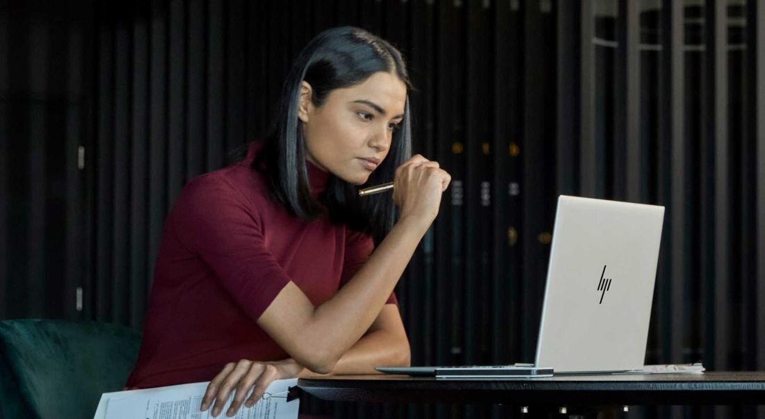 Kobieta w bordowej bluzce koncentrująca się podczas pracy na laptopie HP EliteBook 665 G11, trzyma długopis przy ustach w geście zastanowienia, w tle wnętrze biurowe z metalowymi elementami. - Srebrna elegancka obudowa, wymiary   