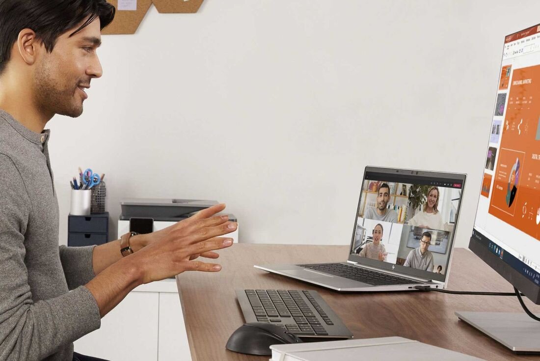 Mężczyzna w szarej bluzie gestykulujący podczas wideokonferencji na laptopie HP EliteBook 665 G11, na ekranie cztery osoby w oknach rozmowy wideo, biuro z monitorem w tle. - Opis serii EliteBook 665 G11 