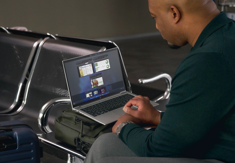 Laptop HP EliteBook 645 G11 Mężczyzna w ciemnej koszuli siedzi na metalowej ławce na lotnisku, korzystając z laptopa o srebrnej obudowie, który leży na torbie. Na ekranie otwarte są kilka okien z aplikacjami, opis