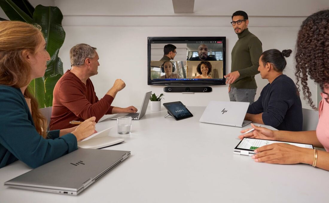 Grupa osób podczas spotkania biznesowego w sali konferencyjnej, na środku stołu leży kilka laptopów HP, na ścianie duży ekran prezentujący wideokonferencję z czterema uczestnikami, na stole tablet sterujący i szklanki z wodą, po lewej duża roślina doniczkowa. - Technologia HP Sure Click