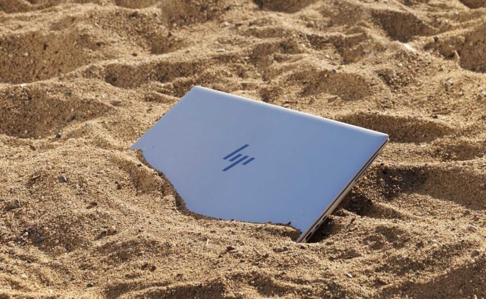 Zamknięty srebrny laptop HP EliteBook częściowo zakopany w piasku na plaży, widoczne charakterystyczne logo HP na pokrywie, wokół drobne kamyczki i ziarenka piasku. - certyfikaty ENERGY STAR i TCO