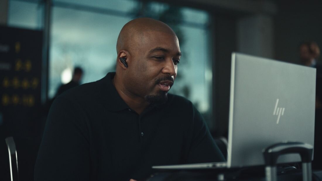 Mężczyzna w ciemnej koszuli pracuje na otwartym laptopie HP EliteBook 845 w przestrzeni publicznej, laptop ustawiony na walizce podróżnej, w tle rozmyte wnętrze budynku z dużymi oknami. - procesor AMD Ryzen