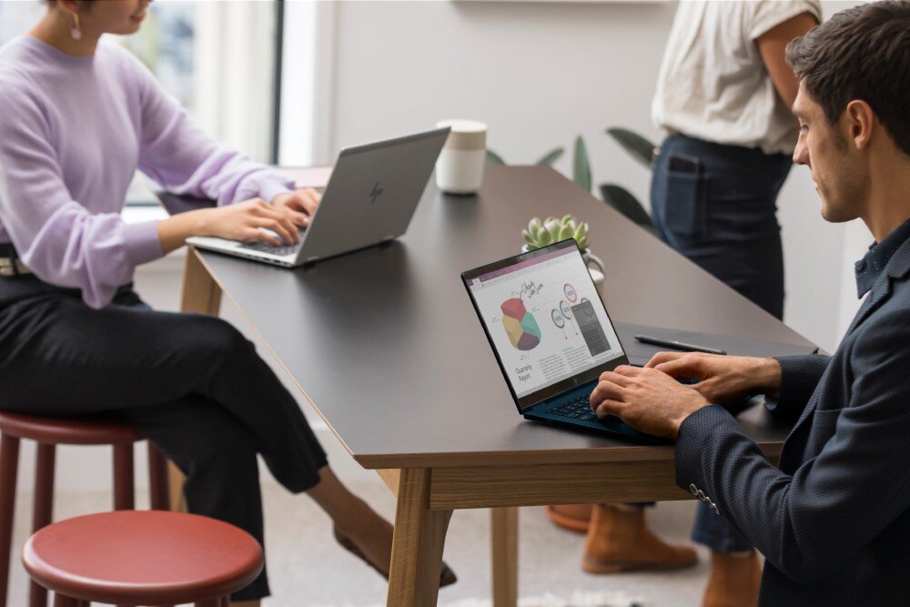 Laptop HP EliteBook 645 G11 Mężczyzna w ciemnym ubraniu siedzi przy drewnianym stole w biurze, korzystając z laptopa. W tle kobieta w jasnofioletowym swetrze również pracuje na laptopie, pamięć RAM, taktowanie, dysk, pojemność