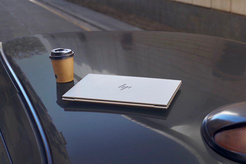 Laptop HP EliteBook 645 G11 Zamknięty laptop o srebrnej obudowie leży na karoserii samochodu. Obok znajduje się papierowy kubek z kawą, bateria, ładowanie