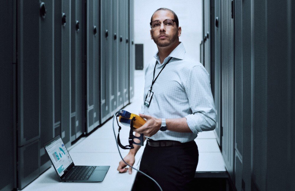 Laptop HP Elitebook 640 G11 Mężczyzna w białej koszuli i okularach znajduje się w serwerowni, trzymając w ręku urządzenie diagnostyczne. Obok na stole stoi otwarty laptop wyświetlający wykresy, zabezpieczenia, funkcje, czytnik linii papilarnych