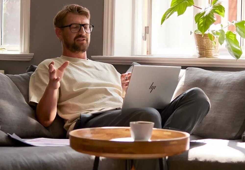 Laptop HP Elitebook 640 G11 Mężczyzna w beżowej koszulce i okularach siedzi na kanapie w salonie, rozmawiając przez wideokonferencję na laptopie, głośniki, dźwięk, mikrofony