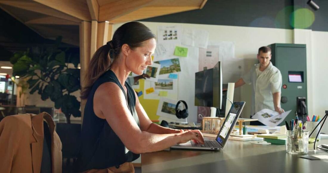 Kobieta pracująca przy biurku na laptopie HP Elite x360 1040 G11, biurowe otoczenie z monitorami, notatkami i roślinami, ekran laptopa wyświetla aplikacje biurowe, w tle mężczyzna z dokumentami. - kamera IR 5 Mpx z automatycznym kadrowaniem