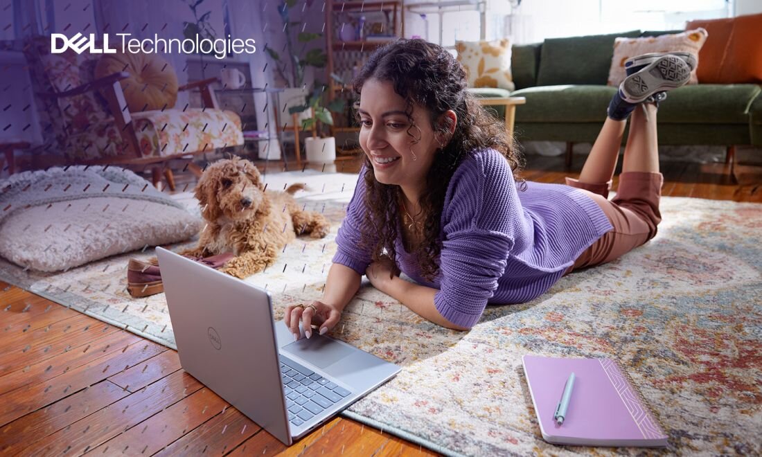 Osoba leżąca na dywanie w salonie, pracująca na laptopie Dell Inspiron 14 Plus 7441, obok pies, notes i długopis, wnętrze z zieloną sofą i poduszkami, logo Dell Technologies w lewym górnym rogu. 