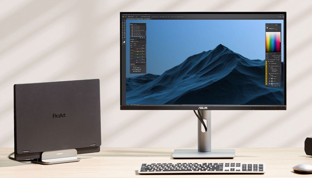 Monitor ASUS ProArt PA278CFRV Monitor na biurku z wyświetlonym ciemnym zdjęciem gór oraz otwartym edytorem graficznym. Obok stoi zamknięty laptop z logo ProArt, powłoka LuxPixel, technologia antyodblaskowa