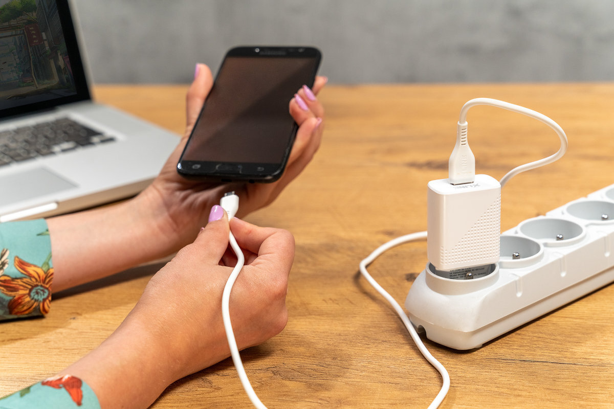 Osoba trzyma smartfon i podłącza go do ładowarki sieciowej umieszczonej w listwie zasilającej na drewnianym stole. W tle widoczny jest otwarty laptop.