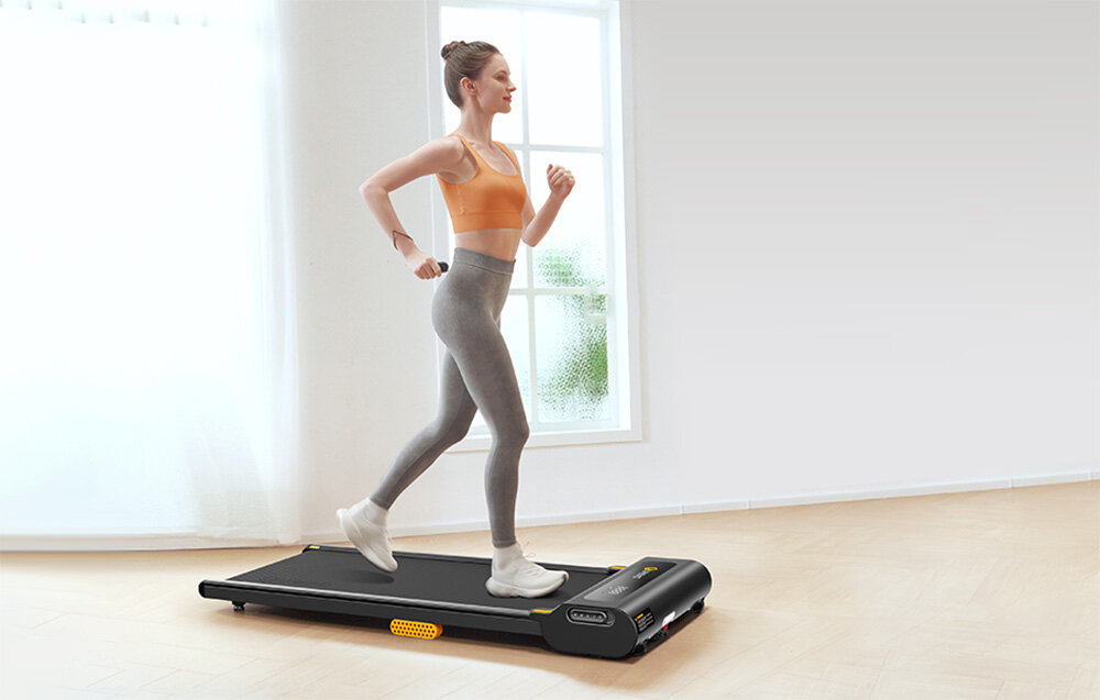 Bieżnia elektryczna UREVO Spacewalk SP1 Lite 1677W 6km-h pokój osoba korzysta z bieżni w tle okno biała ściana można ćwiczyć w domu osiem silikonowych tłumików dwa miękkie poduszki ochrona kolana i kostek antypoślizgowy, pięciowarstwowy pas biegowy