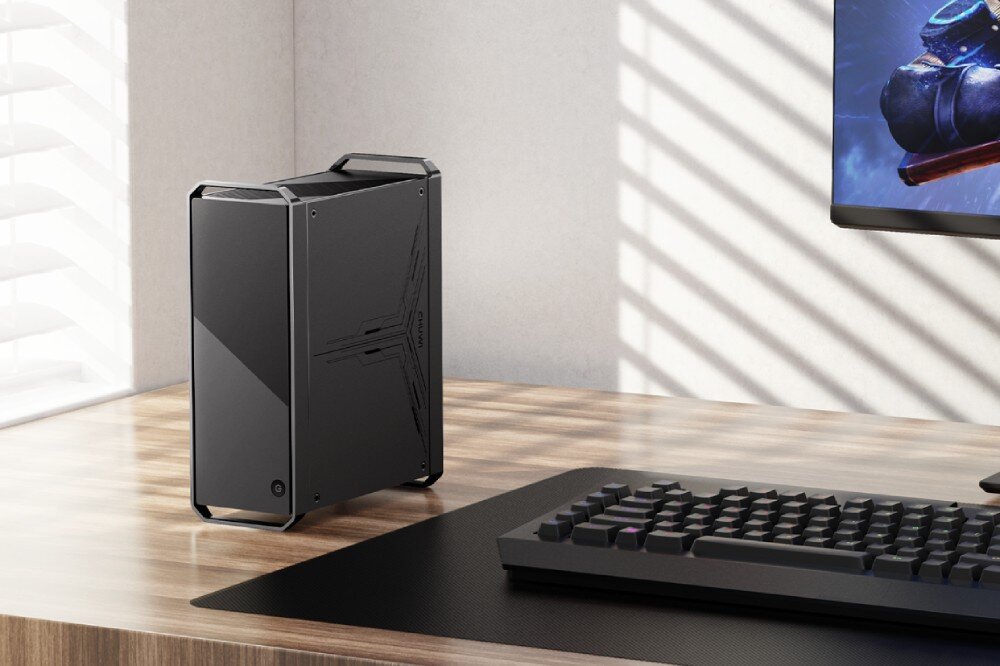 Komputer CHUWI CoreBox CWI601K2 Czarny komputer stojąca pionowo na biurku, z metalowymi uchwytami w rogach. W tle klawiatura i fragment monitora, opis