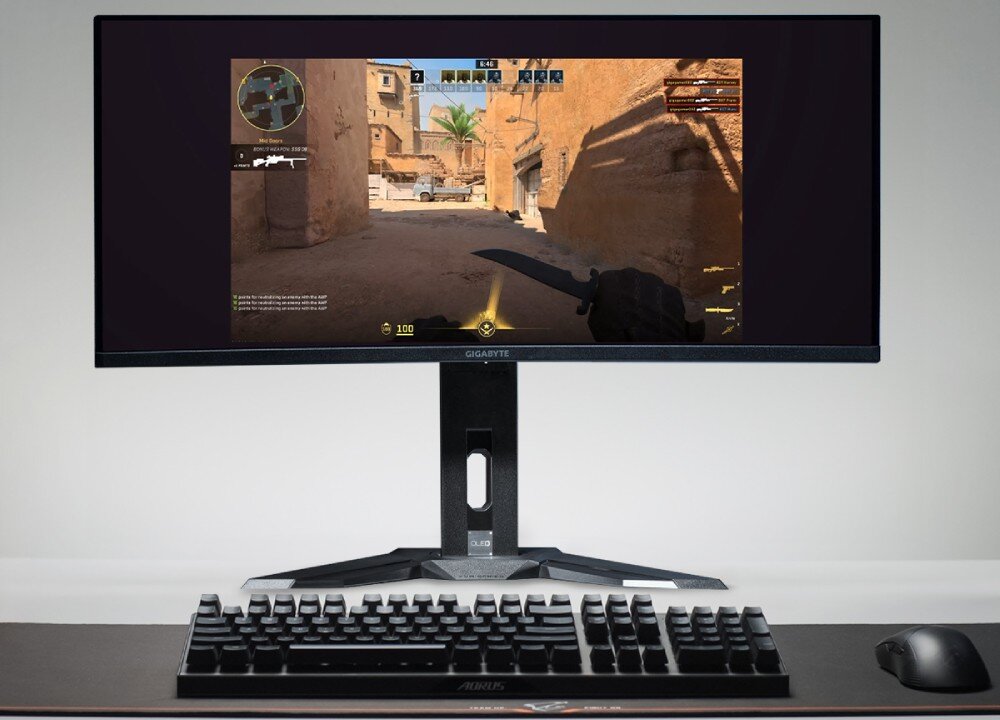 Monitor GIGABYTE MO34WQC Monitor na biurku z uruchomioną grą FPS, przed nim klawiatura i myszka, funkcje, rozwiązania, przełącznik