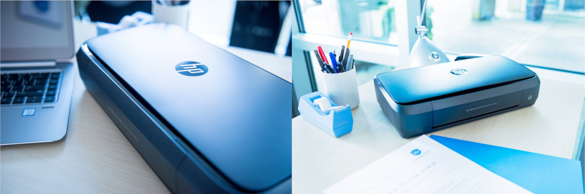 Na biurku znajduje się przenośna drukarka z logo HP, obok laptopa i przyborów biurowych. Drukarka jest w kolorze ciemnym, a na biurku leżą dokumenty i długopisy w kubku.