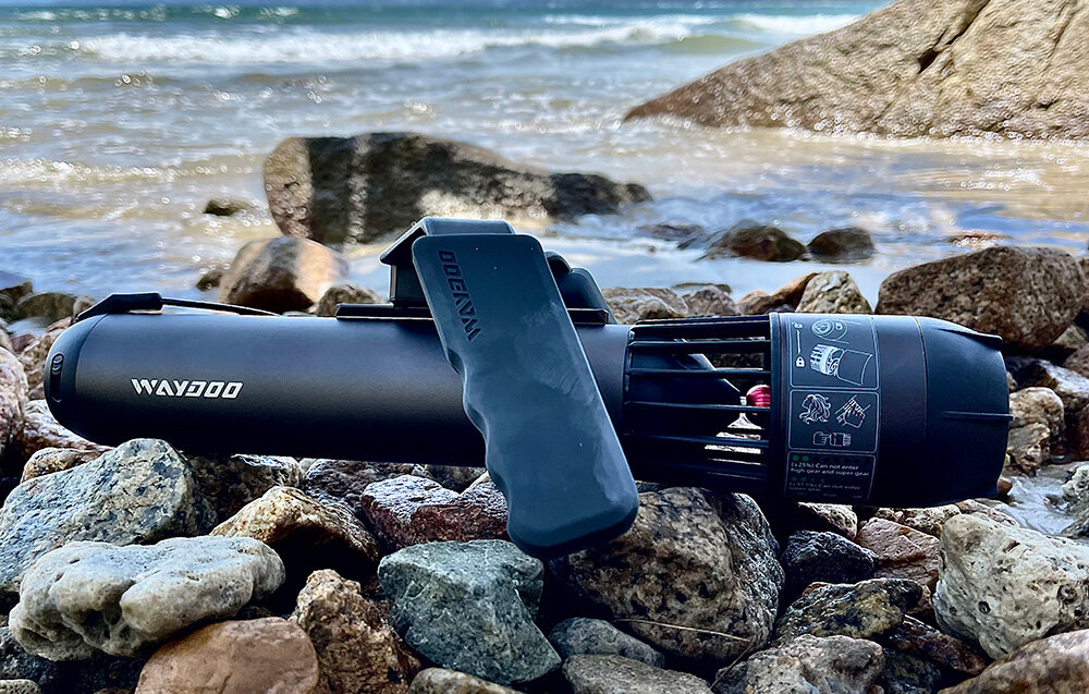 Skuter podwodny WAYDOO Subnado Czarny + zestaw dla SUP plaża kamienie skuter leży na kamieniach w tle woda kontrola komfort szybkość swoboda w wodzie