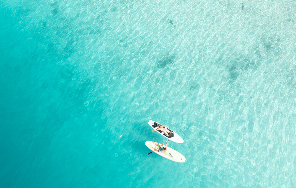 Skuter podwodny WAYDOO Subnado Czarny + zestaw dla SUP woda plaża dwie osoby na supach pływają po wodzie widok z góry waga około 1,4 kg wymiary wynoszą 38 x 6 cm poręczny akumulator o pojemności 98 Wh przewożenie urządzenia na pokładzie samolotu