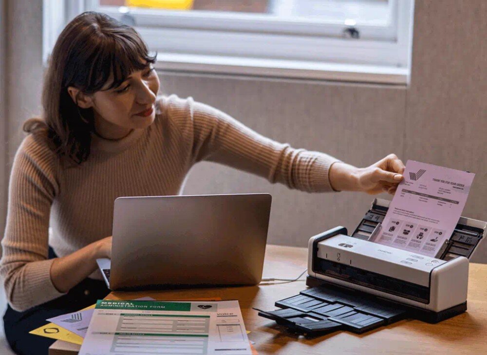 Skaner BROTHER ADS-1300 Kobieta siedząca przy biurku wkłada dokument do skanera. Obok znajduje się laptop i kilka arkuszy papieru, opis