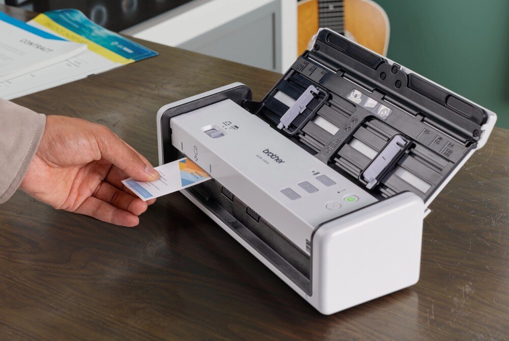 Skaner BROTHER ADS-1300 Zbliżenie na rękę wsuwającą plastikową kartę do dedykowanego gniazda w skanerze. Urządzenie znajduje się na drewnianym blacie, a obok leżą dokumenty, automatyczny podajnik, skanowanie kart