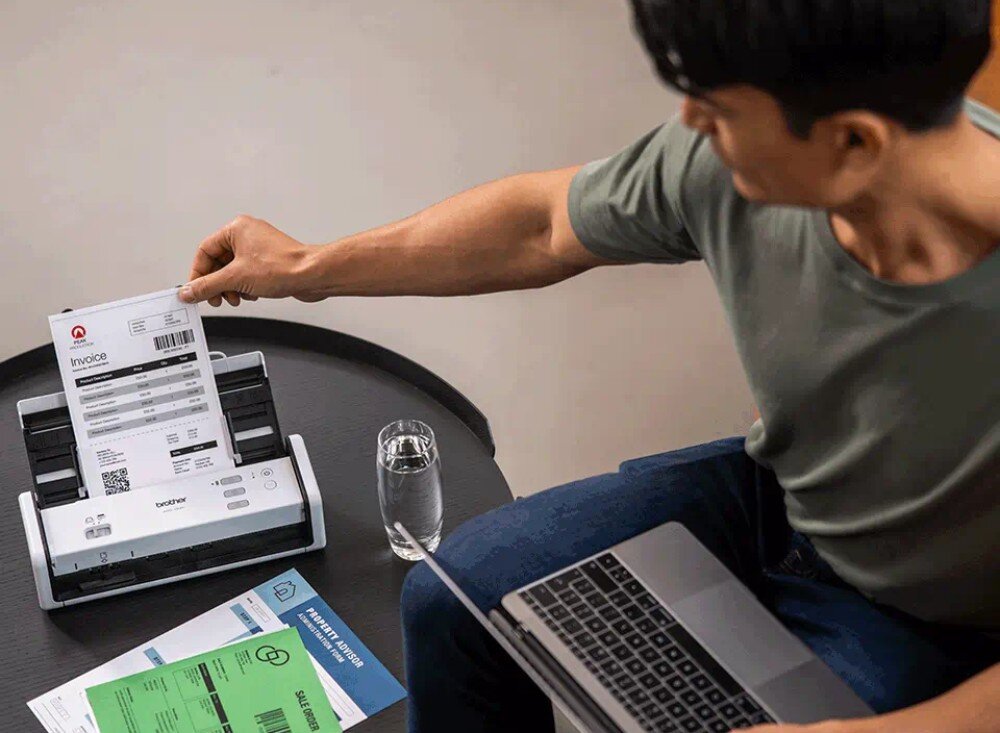 Skaner BROTHER ADS-1300 Mężczyzna siedzący przy okrągłym stoliku, skanujący dokument z fakturą. W drugiej ręce trzyma laptopa, a obok znajduje się szklanka wody i kilka arkuszy papieru, skróty, oprogramowanie, funkcje