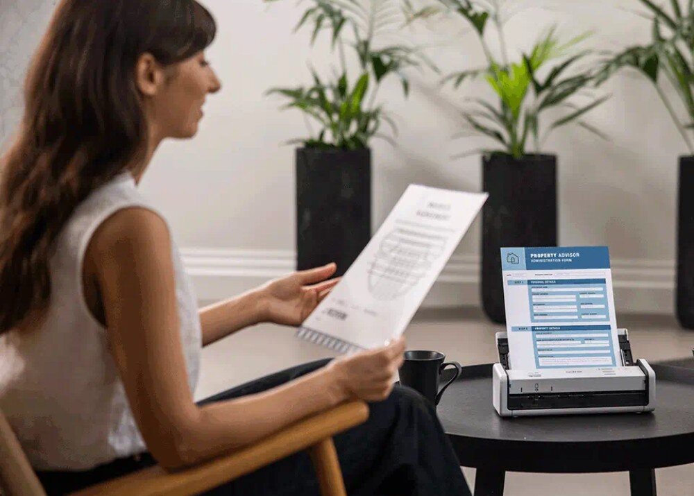 Skaner BROTHER ADS-1300 Kobieta w białej koszuli siedzi na fotelu, trzymając wydrukowany dokument i porównując go z arkuszem skanowanym przez urządzenie, rozdzielczość, technologia LED, sensor