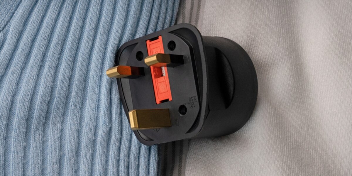 Czarny adapter podróżny z trzema metalowymi bolcami i czerwonym elementem na środku. Adapter leży na tkaninie w kolorze niebieskim i szarym.