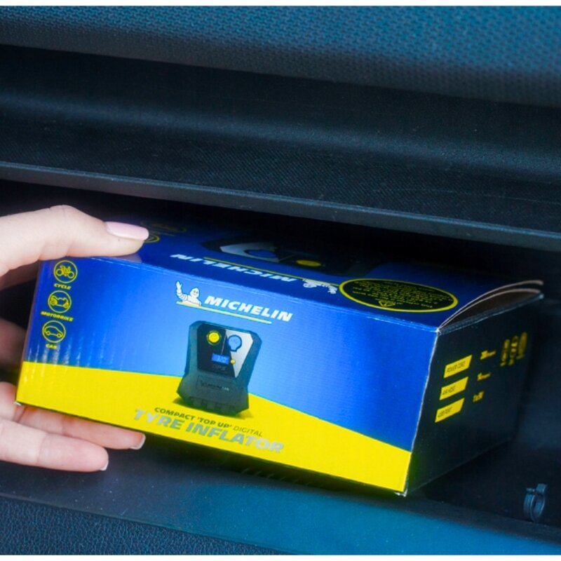 Osoba trzyma pudełko z napisem \'MICHELIN COMPACT TOP UP DIGITAL TYRE INFLATOR\' w kolorach niebieskim i żółtym. Na pudełku widoczne są ikony roweru i samochodu oraz logo Michelin.