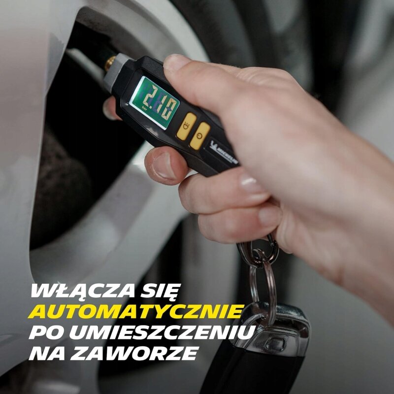 Osoba trzyma manometr ciśnieniomierz przy zaworze koła samochodowego, na wyświetlaczu widoczny jest wynik 2.10. Tekst na obrazie: \'WŁĄCZA SIĘ AUTOMATYCZNIE PO UMIESZCZENIU NA ZAWORZE\'.