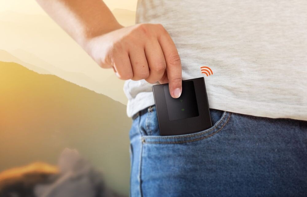 Router TENDA 4G180 v4.0 Osoba w jasnym t-shircie i jeansach wkłada mały, czarny, prostokątny ruter do przedniej kieszeni spodni na tle rozmytego krajobrazu górskiego o zachodzie słońca