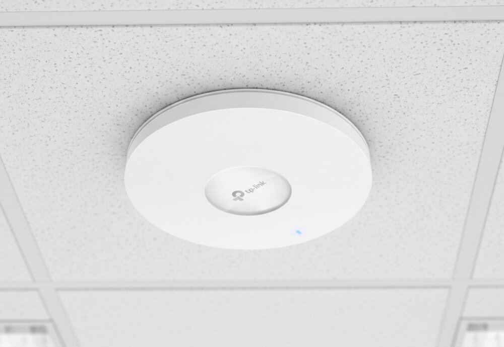 Na obrazie widoczny jest biały punkt dostępu zamontowany na suficie z logo \'tp-link\' na środku. Urządzenie ma okrągły kształt i świeci się na nim niebieska dioda.