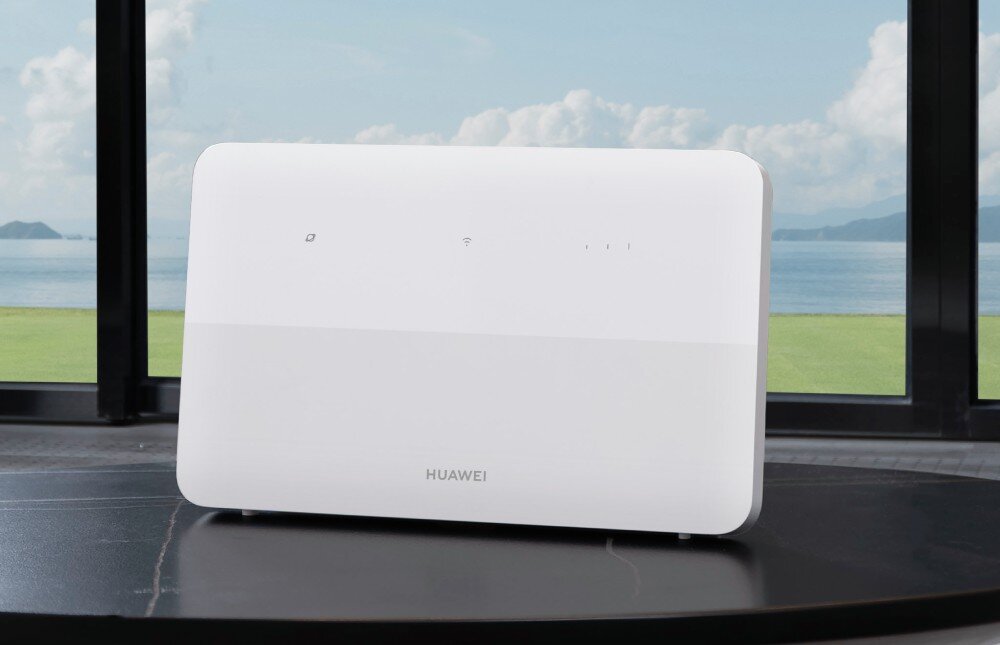 Router HUAWEI 4G CPE 5 B636-336 Biały router o prostokątnym kształcie z zaokrąglonymi rogami i logo Huawei stoi na czarnym stole na tle okna z widokiem na zieleń i wodę