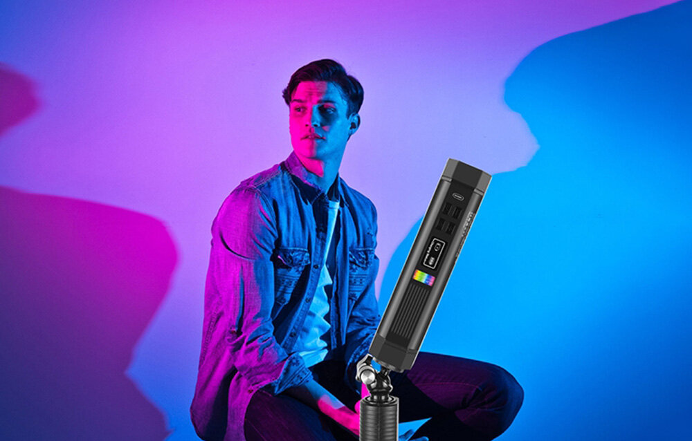 Mężczyzna siedzi na tle kolorowego oświetlenia, a obok niego ustawiona jest lampa fotograficzna LED na statywie.