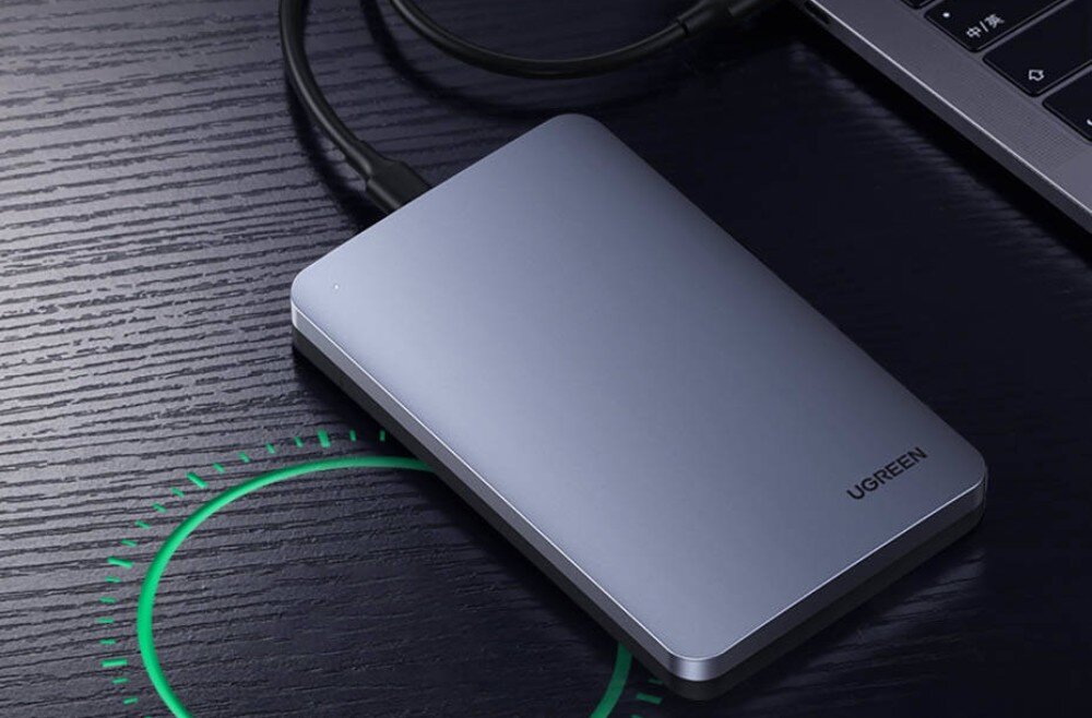 Obudowa dysku UGREEN CM300 Srebrna, prostokątna obudowa z logo Ugreen leżąca na ciemnym, drewnianym blacie obok laptopa. Pod urządzeniem widoczna jest zielona, kolista grafika sugerująca działanie funkcji bezprzewodowych