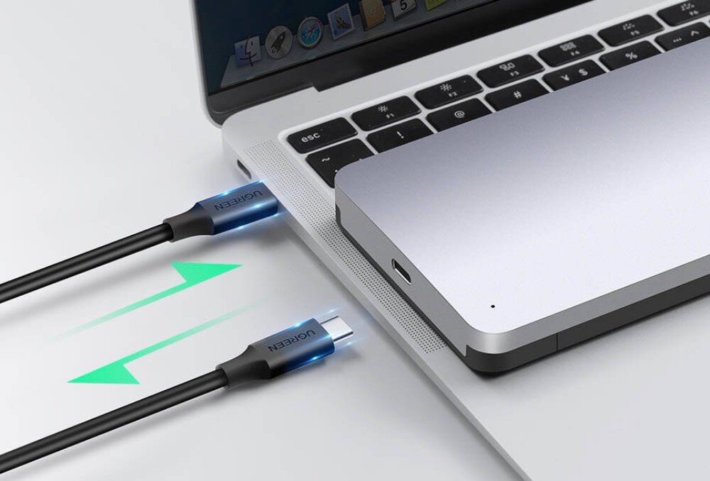 Obudowa dysku UGREEN CM300 Srebrna obudowa leżąca na obudowie laptopa. Obok widoczne są dwa kable USB-C skierowane w stronę portów, a między nimi zielona strzałka sugerująca dwukierunkowy przesył danych