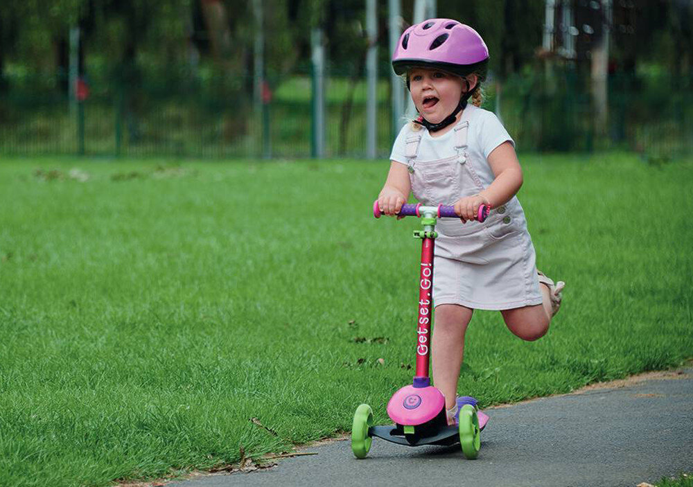 Hulajnoga dla dzieci TRUNKI Mała Różowy park droga dziecko w kasku jedzie na hulajnodze po drodze w tle drzewa trawnik dla dzieci od 3. roku życia rozwój koordynacji i równowagi szeroka platforma stabilna konstrukcja do bezpiecznego balansowania regulowana wysokość kierownicy do rekreacyjnej jazdy uczy aktywności i samodzielności w bezpieczny sposób
