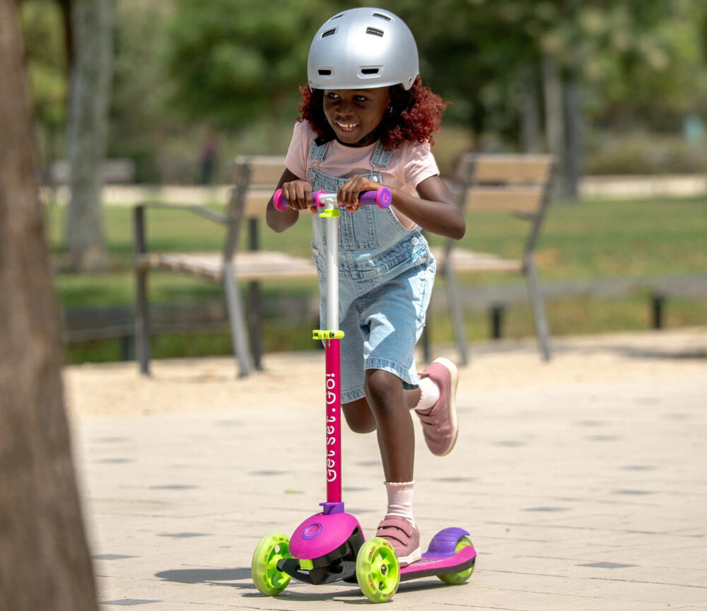 Hulajnoga dla dzieci TRUNKI Mała Różowy park plac zabaw droga dziecko jedzie na hulajnodze w tle drzewa ławki trawnik tylny hamulec nożny prosta i skuteczna kontrola nad prędkością jazdy lekko nacisnąć hamulec uczy odpowiedzialności i przewidywania szerokie tylne koło stabilizuje tor jazdy
