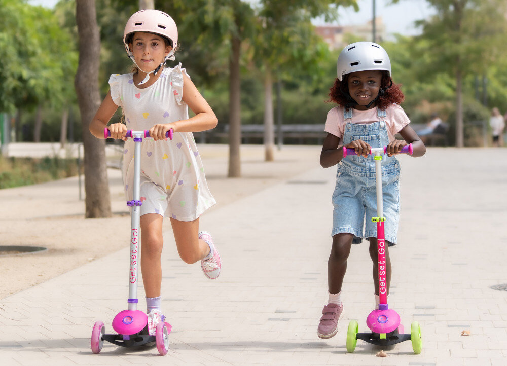 Hulajnoga dla dzieci TRUNKI Mała Różowy park droga dwójka dzieci jedzie na hulajnogach pod drodze w tle drzewa niebo platforma wykonana z polipropylenu i włókna szklanego solidne podparcie stóp dziecka szerokość 12 cm stabilność odporność na uszkodzenia ułatwia naukę jazd sprzyja nauce równowagi