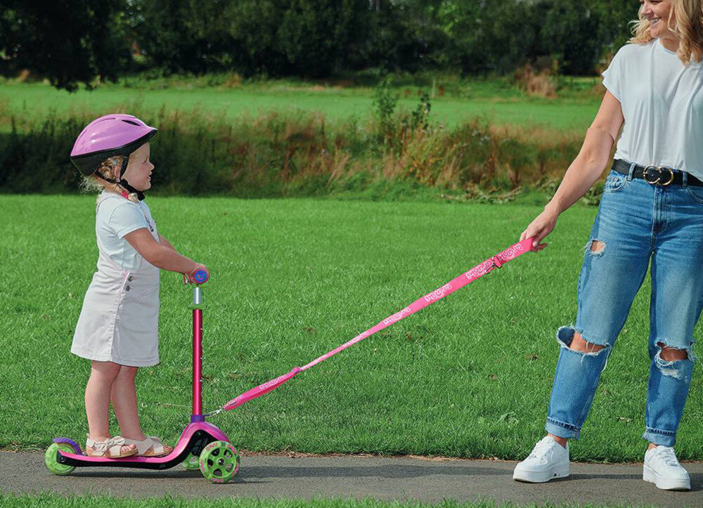 Hulajnoga dla dzieci TRUNKI Mała Różowy park dziecko na hulajnodze osoba trzyma pasek w dłoni ciągnie za sobą hulajnogę w tle trawnik wspiera rozwój mięśni równowagi koordynacji zdrowe spędzanie czasu na świeżym powietrzu bawi uczy i rozwija bezpieczeństwo i wygodę pasek i torba w zestawie rodzic może przenieść hulajnogę lub ciągnąć dziecko za sobą