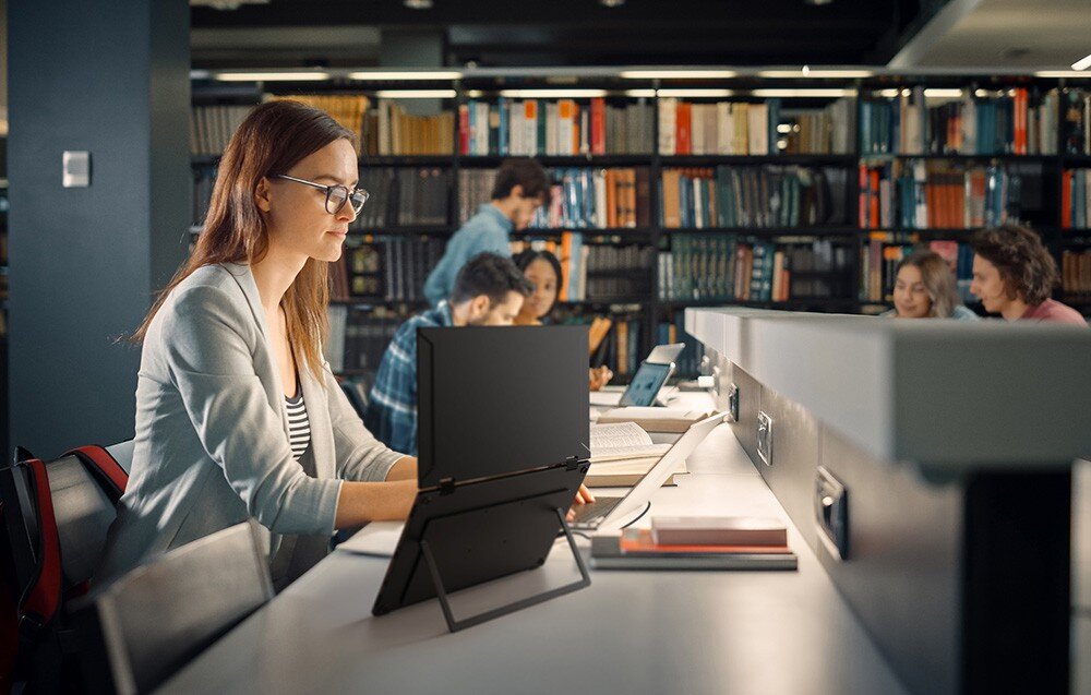 Monitor UPERFECT Ustation Delta Kobieta w okularach pracuje w bibliotece przy przenośnym ekranie ustawionym na biurku, obok otwarta książka i plecak, składanie