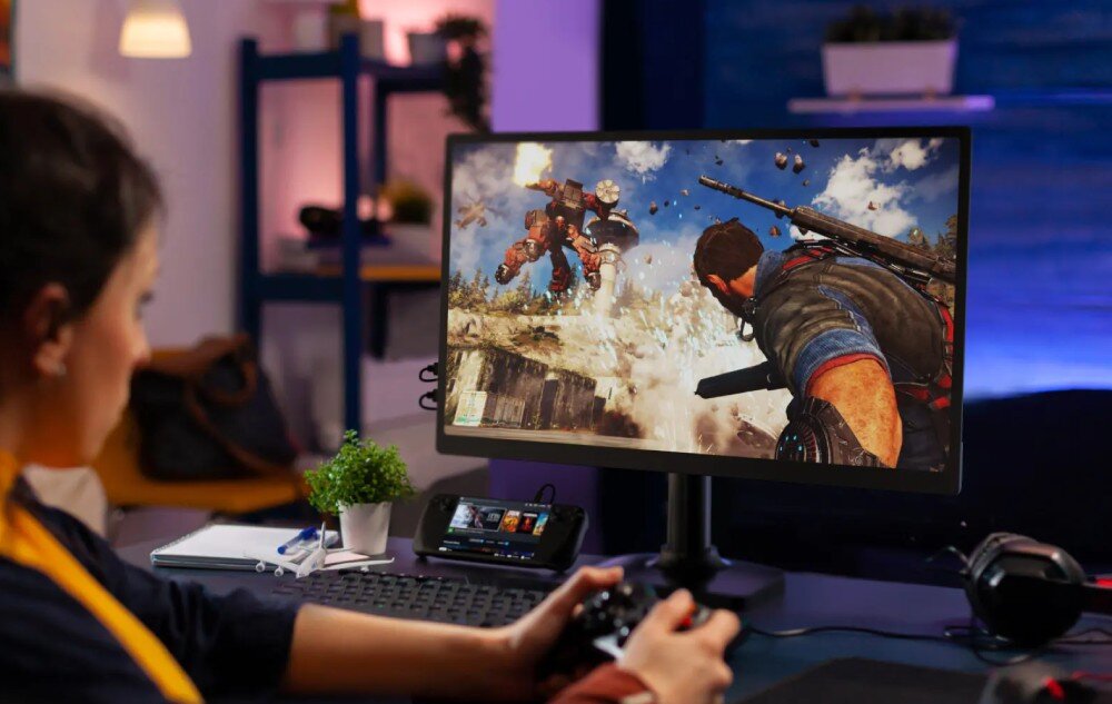 Monitor UPERFECT USteam E6 Pro Osoba grająca na konsoli z kontrolerem w ręku siedzi przed dużym ekranem, na którym wyświetlana jest dynamiczna scena z gry z żołnierzem i robotem bojowym, opis