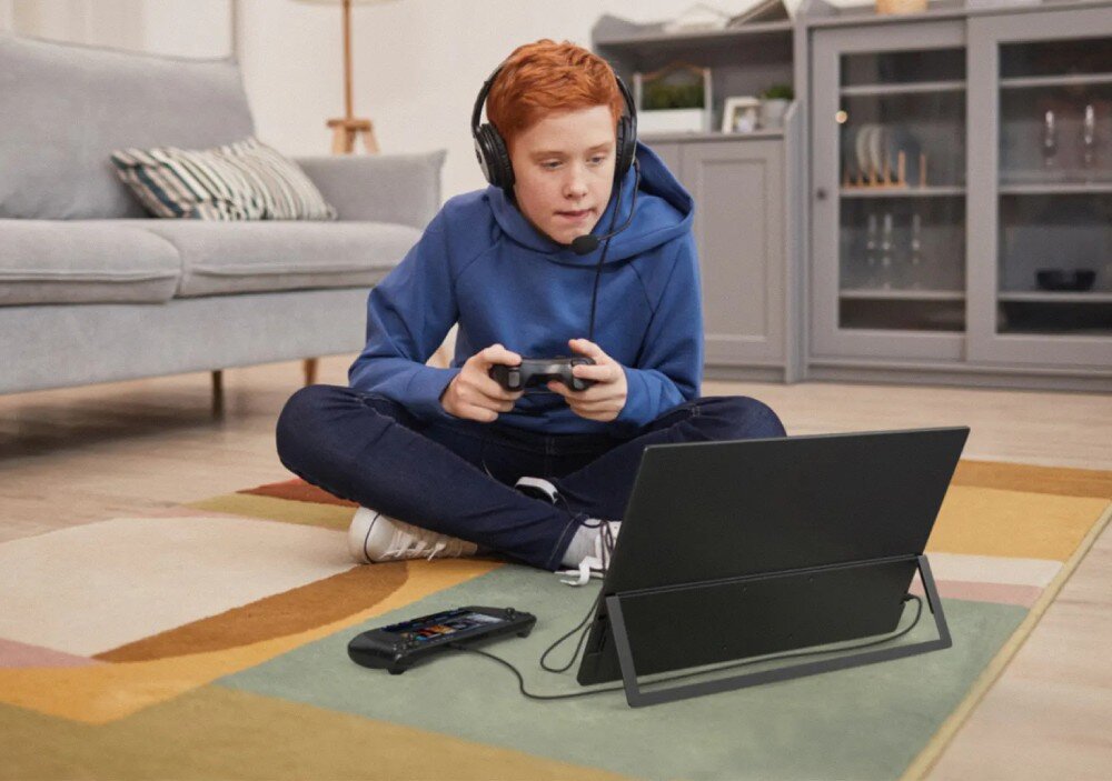 Monitor UPERFECT USteam E6 Pro Chłopiec w słuchawkach z mikrofonem siedzący na dywanie gra na konsoli, przed nim stoi monitor wsparty na wbudowanej nóżce, głośniki, dźwięk, gniazdo słuchawkowe, podstawka, mocowanie VESA