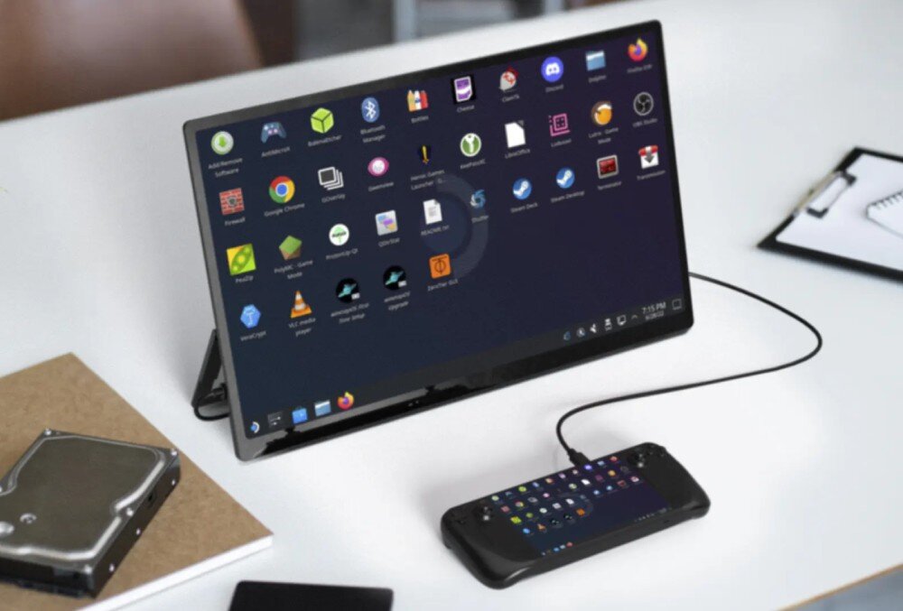Monitor UPERFECT USteam E6 Pro Monitor ustawiony na biurku wyświetlający ekran systemu z ikonami aplikacji, obok na kablu podłączona konsola, w tle leżą notatniki i dysk twardy, zasilanie, porty, podłączenie, kompatybilność
