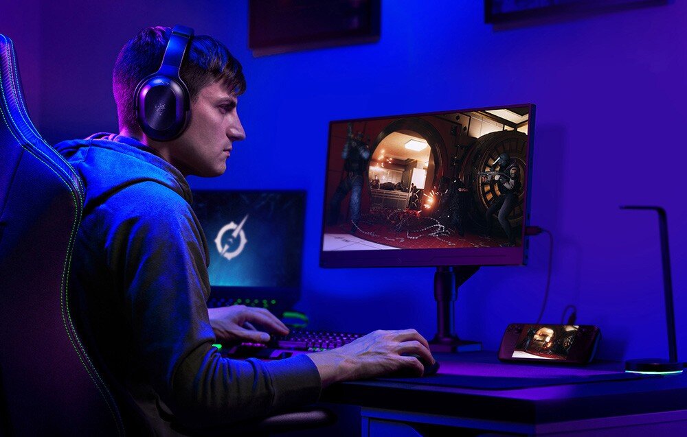 Monitor UPERFECT UGame J5 Mężczyzna siedzący przy biurku w słuchawkach gra na komputerze, przed nim monitor przenośny z wyświetloną sceną strzelaniny, obudowa, waga, ramka, mocowanie VESA