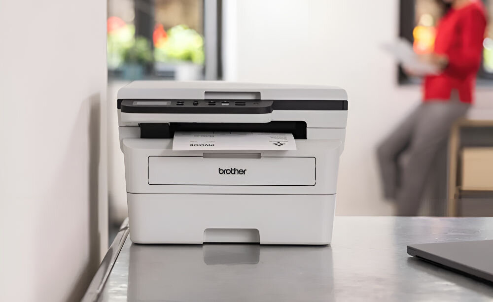 Urządzenie wielofunkcyjne BROTHER DCP-B7620DW Automatyczny druk dwustronny, Wi-Fi prezentacja urządzenia na biurku w tle rozmyte biuro i kobieta rozdzielczość skanowania dokumenty zdjęcia wysoka jakość