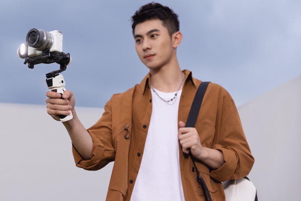 Gimbal ZHIYUN Crane M3S Combo mężczyzna trzymający przed sobą gimbal z zamontowanym aparatem, z plecakiem na ramieniu wygląd opis