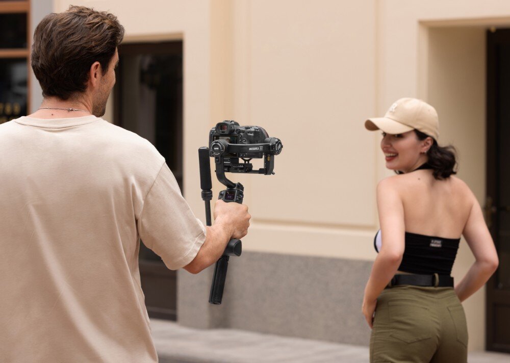 Gimbal ZHIYUN Weebill 3S Mężczyzna filmuje kobietę na ulicy, trzymając stabilizator z aparatem. Kobieta w beżowej czapce i ciemnym topie uśmiecha się do kamery. W tle jasna elewacja budynku, przeznaczenie, kompatybilność, zalety