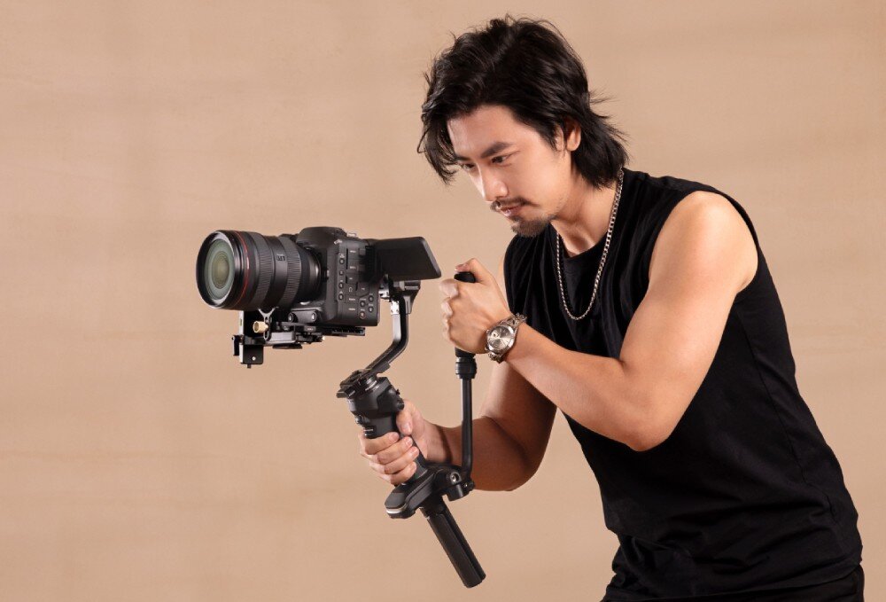 Gimbal ZHIYUN Crane 4 Combo Mężczyzna w czarnym podkoszulku trzyma aparat zamocowany na stabilizatorze, pochylając się lekko do przodu, opis